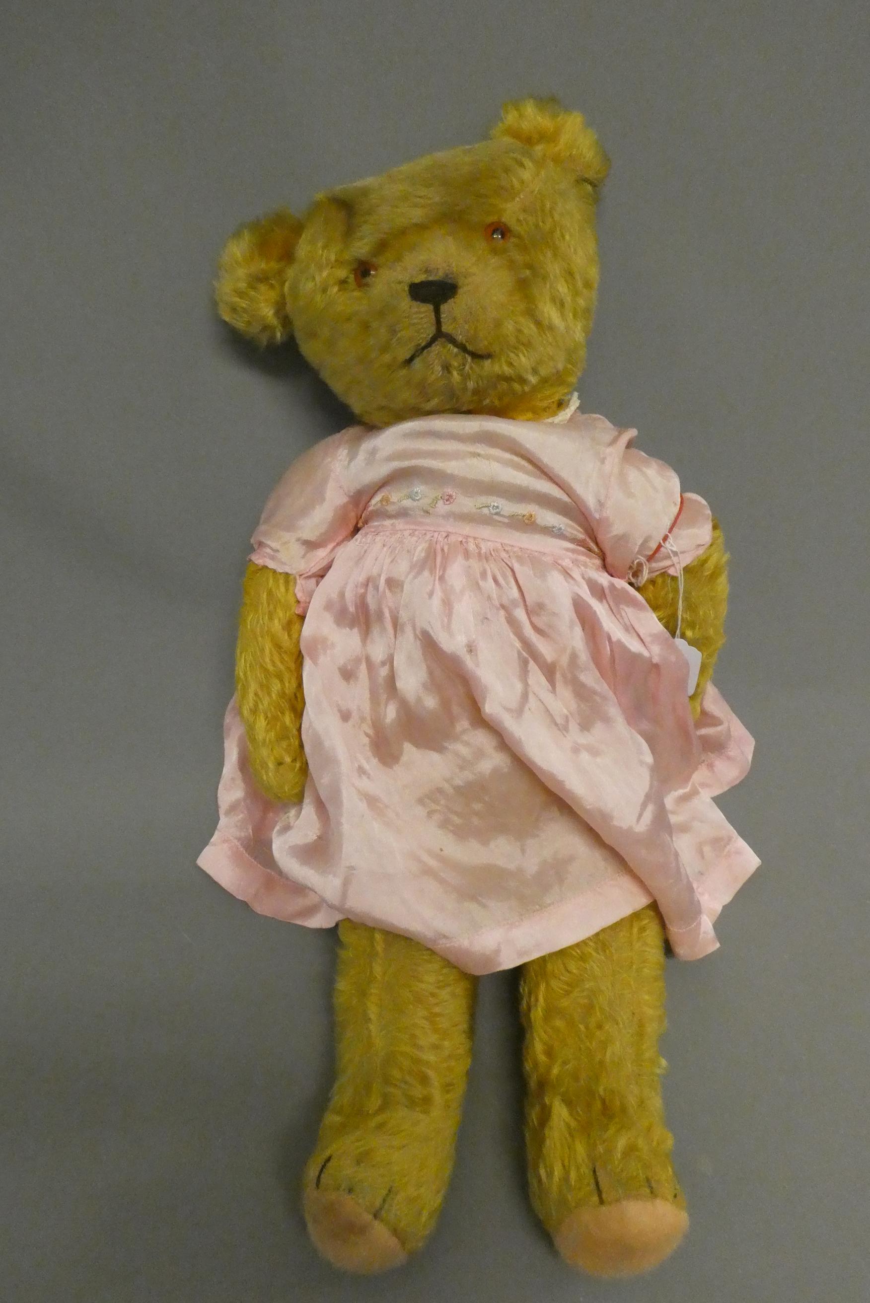 A pre-war teddy bear, with sewn nose, glass amber eyes, blond plush, pink felt pads and pink dress, Image