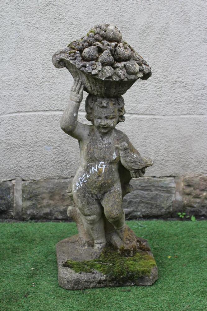 A CAST STONE FIGURE allegorical of Autumn, modelled as a Putto carrying a basket of fruit on his Image