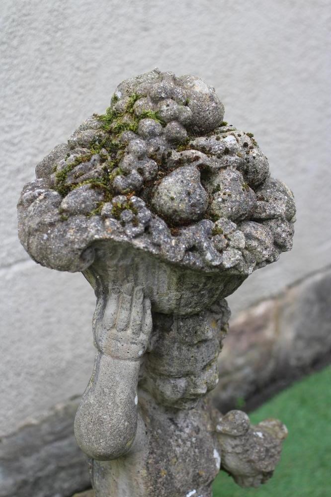 A CAST STONE FIGURE allegorical of Autumn, modelled as a Putto carrying a basket of fruit on his Image