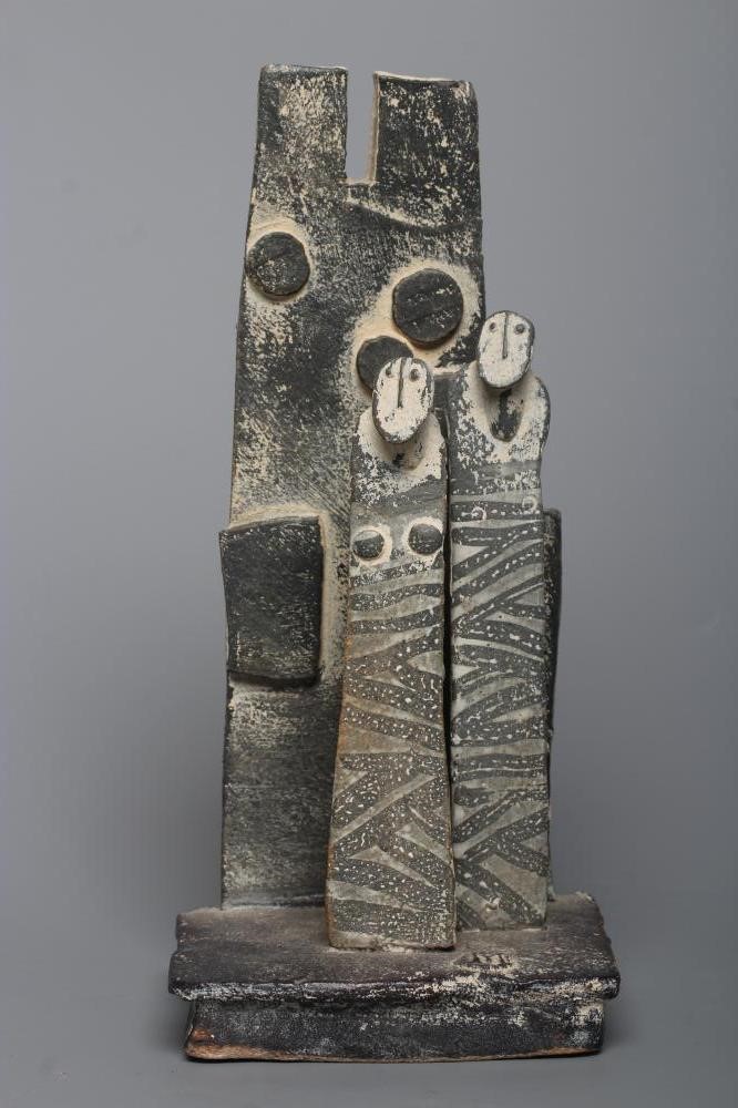 JOHN MALTBY (1936-2000) - a matt grey stoneware sculpture of two figures standing before a cut slab Image