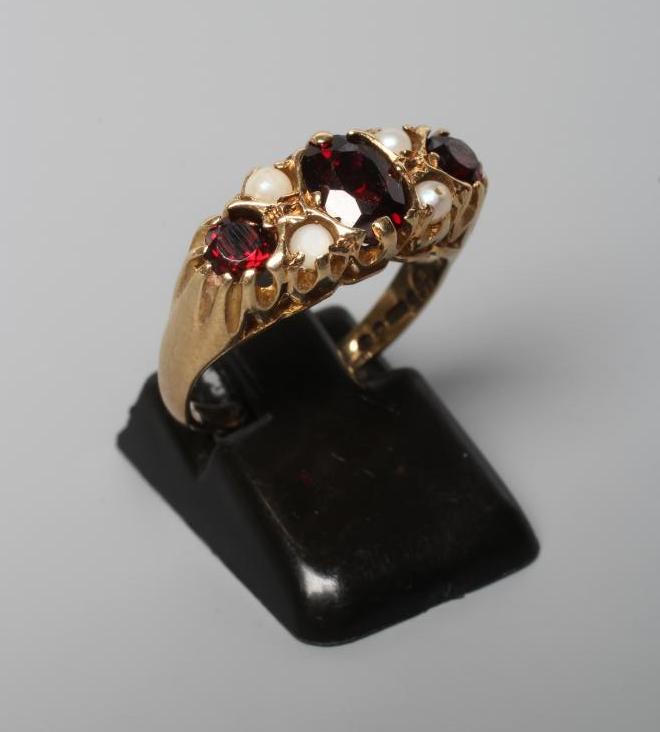 A THREE STONE GARNET RING, the facet cut stones claw set with four seed pearls between, on carved Image