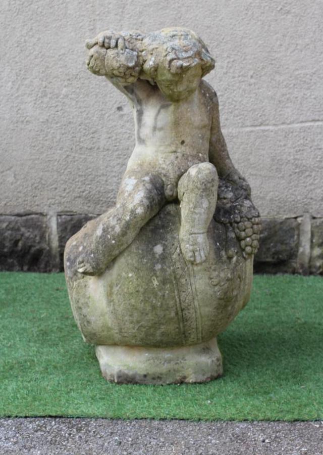 A CAST STONE FIGURE OF A BACCANALIAN PUTTO, modelled seated on an overturned wine jar and holding Image