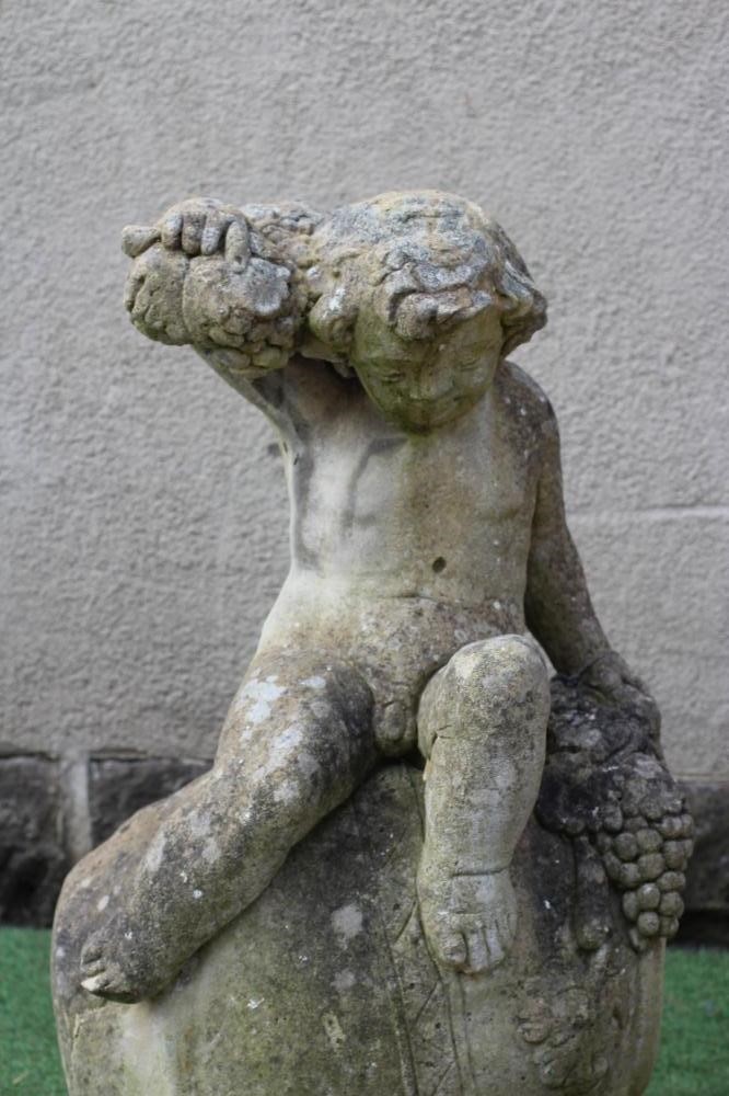 A CAST STONE FIGURE OF A BACCANALIAN PUTTO, modelled seated on an overturned wine jar and holding Image