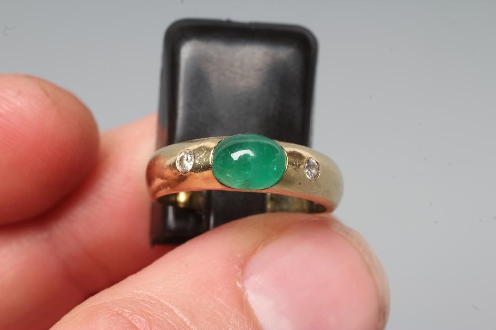 AN EMERALD AND DIAMOND RING, the oval cabochon polished emerald tension set and flanked by two gypsy Image