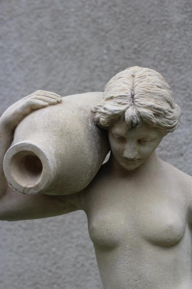 A CAST STONE FIGURE OF A YOUNG MAIDEN modelled semi-nude and holding a water vessel on her shoulder, Image