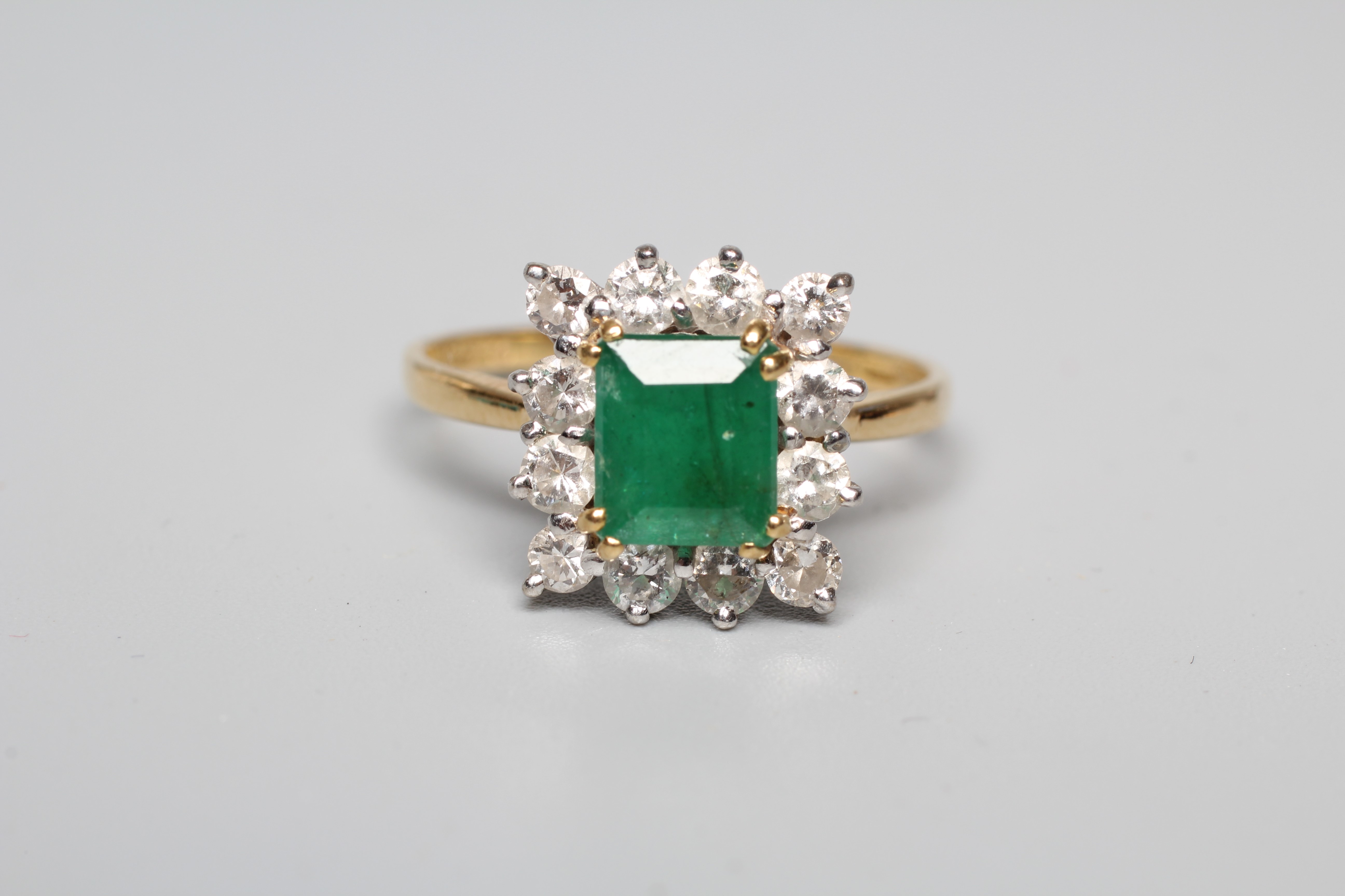 AN EMERALD AND DIAMOND CLUSTER RING, the step cut emerald claw set to a border of twelve round Image