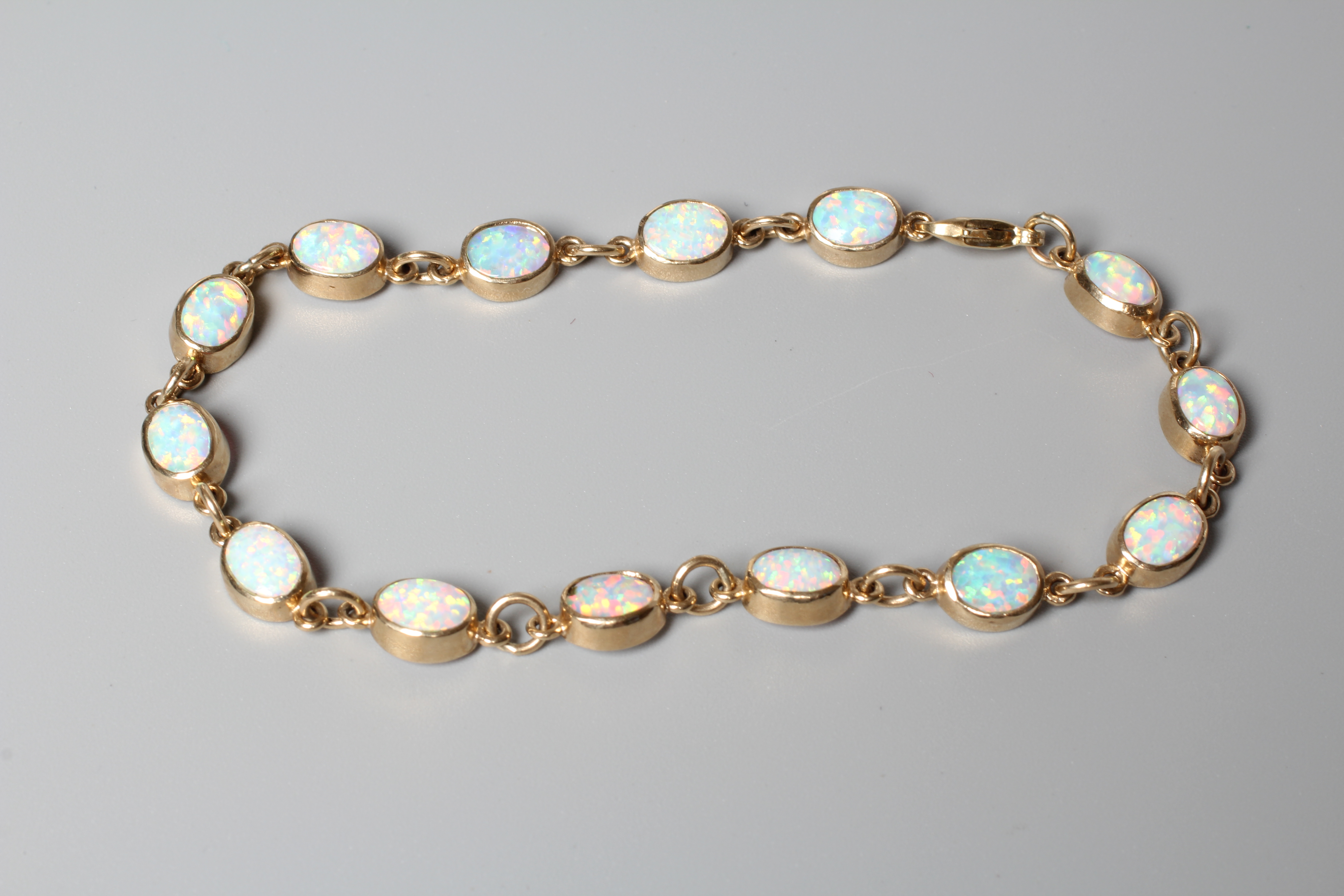 AN OPAL BRACELET, the fourteen oval cabochon polished stones open back collet set to a plain frame Image