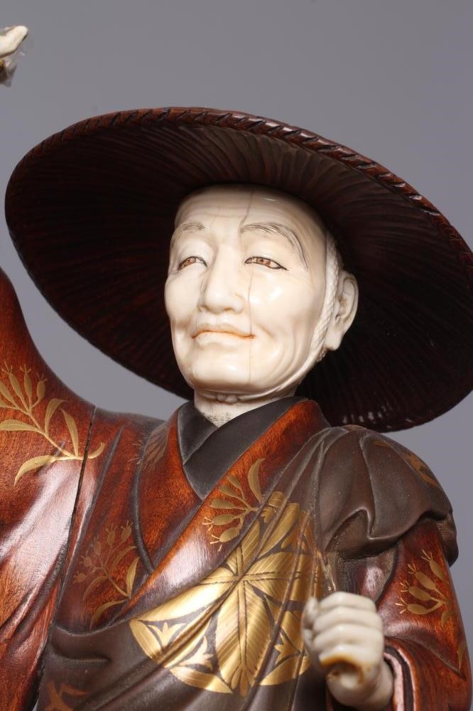 A JAPANESE CARVED WOOD AND IVORY OKIMONO modelled as a man wearing a kasa and gold highlighted Image