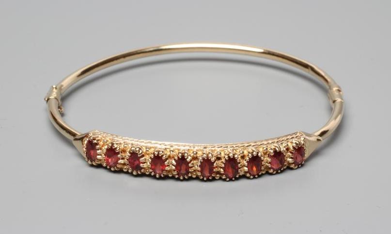 A STIFF HINGED BANGLE, the upper section claw set with ten oval facet cut garnets, indistinctly Image