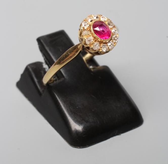 A VICTORIAN RUBY AND DIAMOND CLUSTER RING, the oval facet cut ruby open back collet set to a border Image