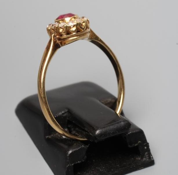 A VICTORIAN RUBY AND DIAMOND CLUSTER RING, the oval facet cut ruby open back collet set to a border Image