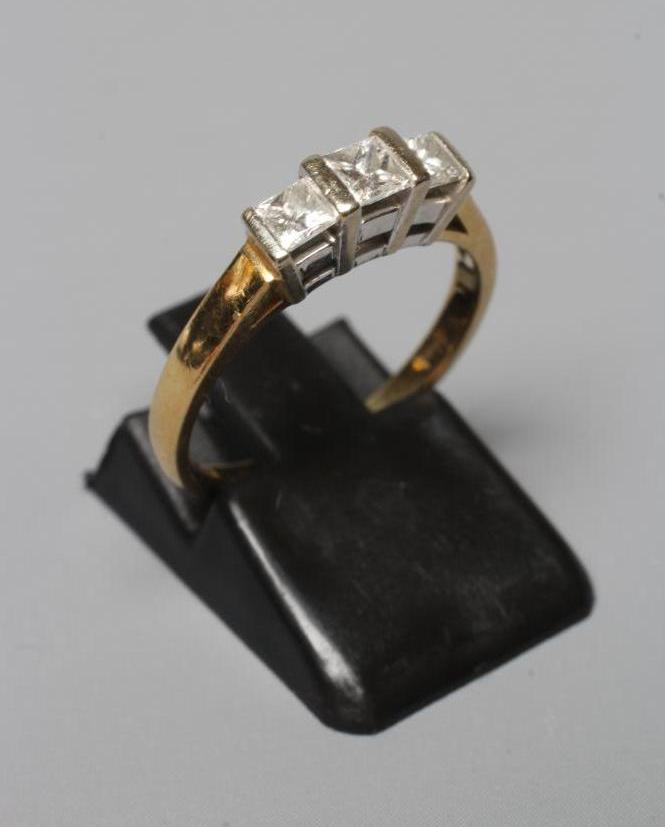 A THREE STONE DIAMOND RING, the Princess cut stones bar set to a plain shank, stamped 18K CORONA, Image