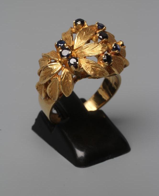 A COCKTAIL RING, the high flowerhead panel claw set with nine small sapphires to a plain wide shank Image