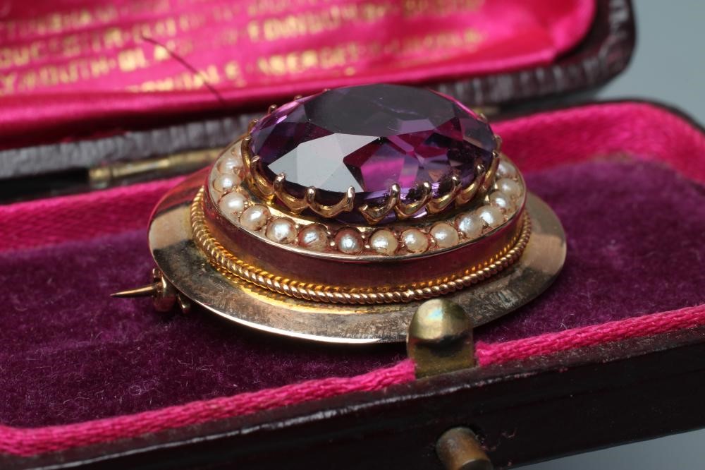 A LATE VICTORIAN AMETHYST BROOCH, the oval facet cut stone set to a border of seed pearls in a plain Image